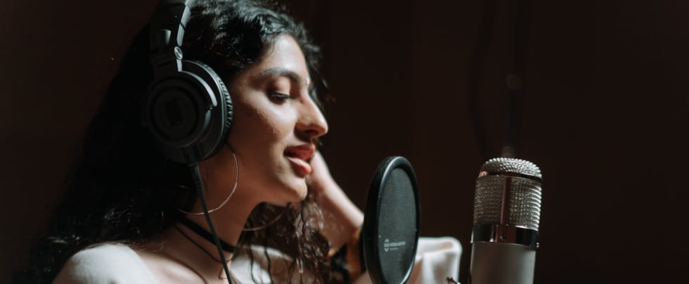 Woman in a music studio singing into a microphone, wearing headphones.